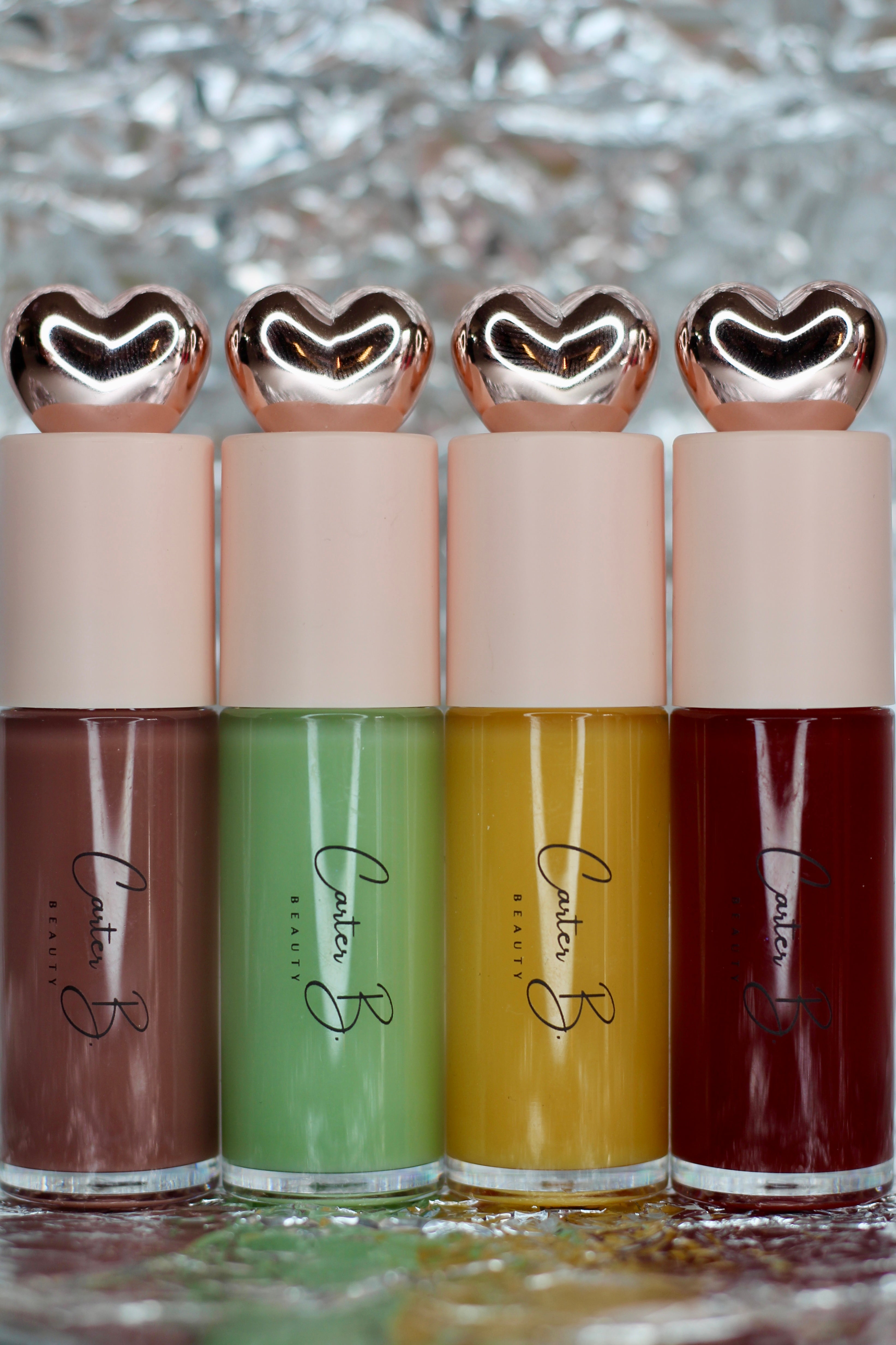 Vier neue Lippenöle von Carter B. Beauty. In einem zarten Braunton (LATTE), Grünton (AVOCADO), Gelbton (GINGER) und Rosenholzton (ROSEWOOD), vor einem silbernden Hintergrund