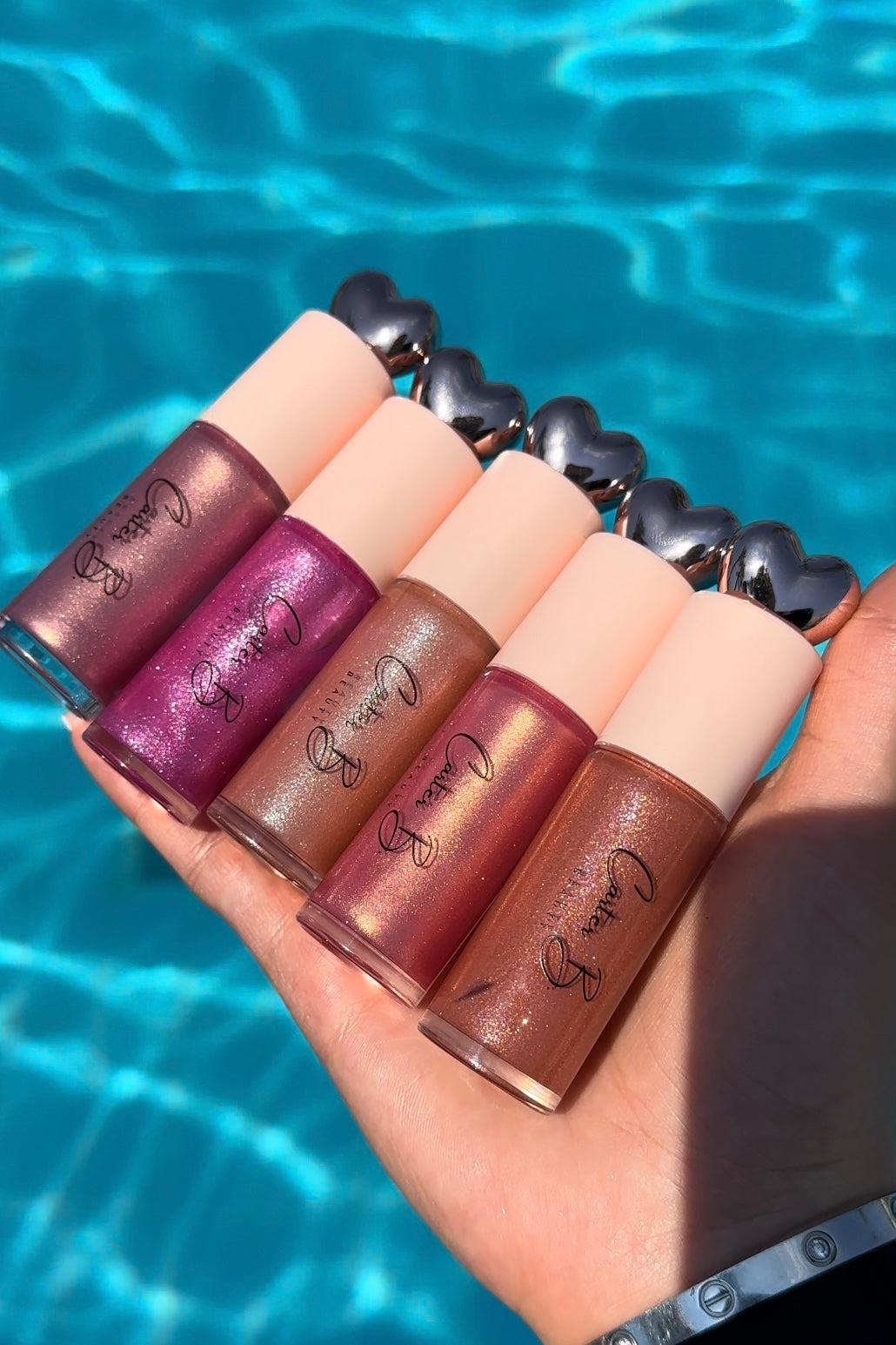 Eine Hand hält fünf Carter B. Beauty Lipgloss in schimmernden Pink-, Lila- und Nude-Tönen vor einem türkisblauen Pool, vegan und tierversuchsfrei für einen funkelnden Sommerlook
